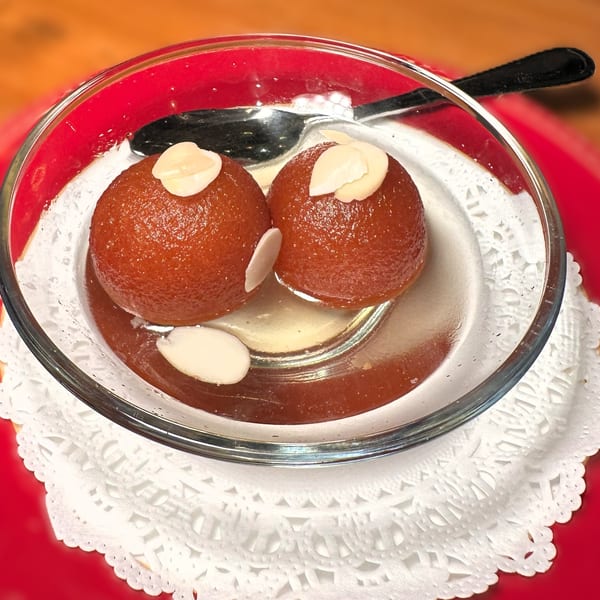 Two gulab jamun, garnished with almond slivers, in a glass bowl with a spoon.