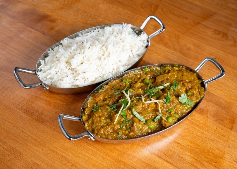Two silver bowls on a wooden table, one with white rice and the other with curry.
