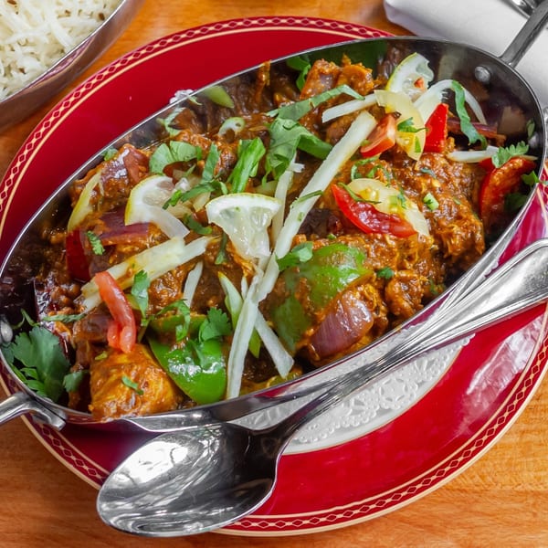 Indian curry in a metal serving dish with rice, garnished with lemon, cilantro, and peppers.
