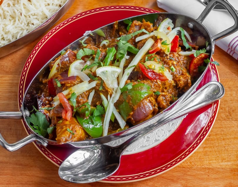 Indian curry in a metal serving dish with rice, garnished with lemon, cilantro, and peppers.