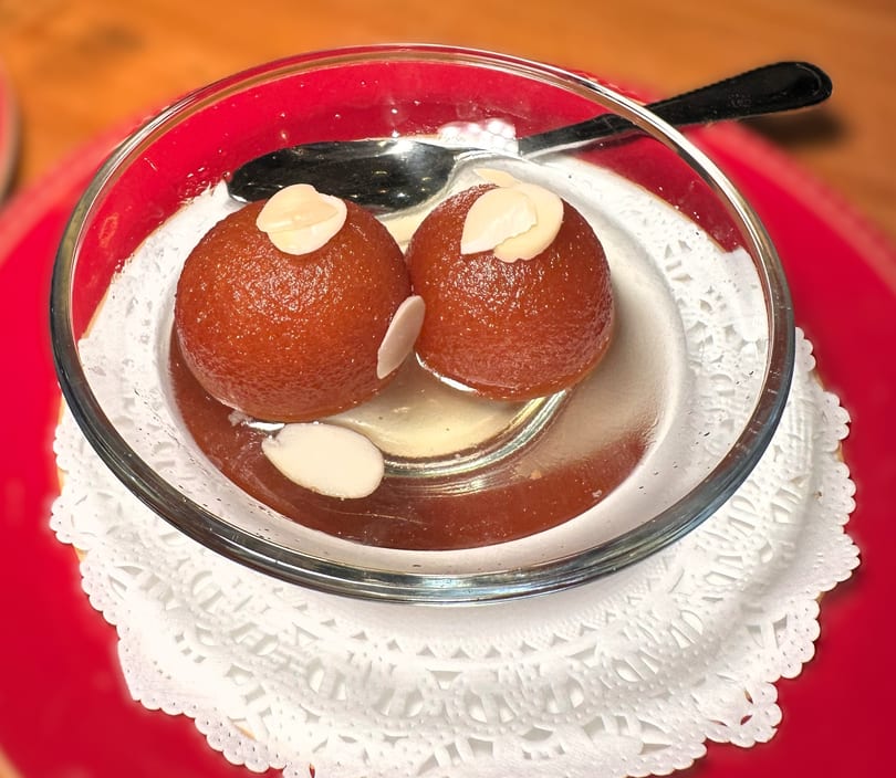 Two gulab jamun with almond slivers in syrup, served in a glass bowl on a white doily.