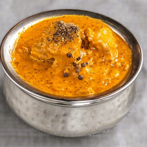 A silver bowl filled with orange curry, topped with black pepper, on a light gray surface.