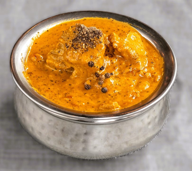 A silver bowl filled with orange curry, topped with black pepper, on a light gray surface.