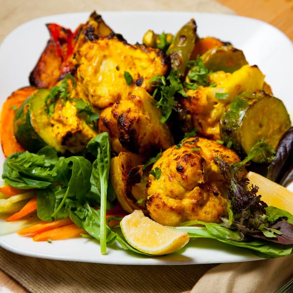 Grilled chicken tikka with roasted vegetables, fresh greens, and a lemon wedge on a white plate.