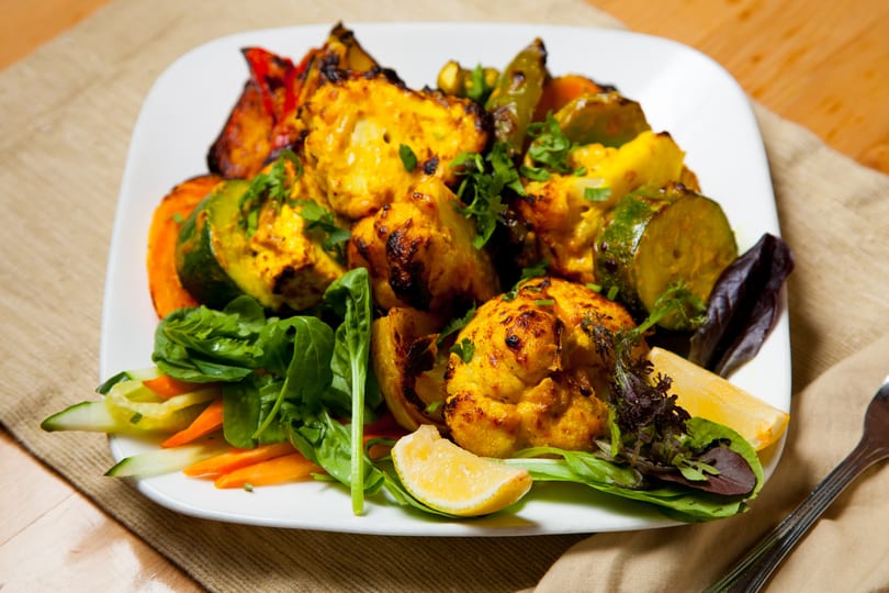Grilled chicken tikka with roasted vegetables, fresh greens, and a lemon wedge on a white plate.