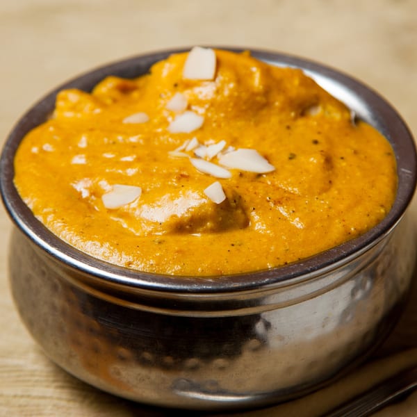 A metal bowl filled with orange curry, garnished with white almond slivers, on a light brown surface.