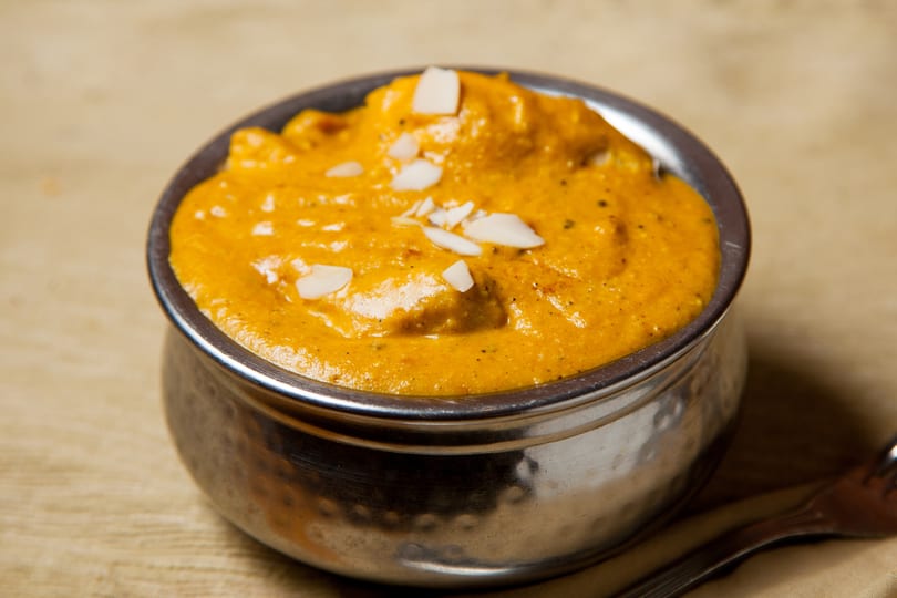 A metal bowl filled with orange curry, garnished with white almond slivers, on a light brown surface.