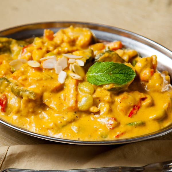 A vibrant yellow curry dish with vegetables, almonds, and a mint leaf in a silver bowl.