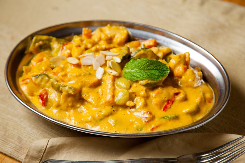 A vibrant yellow curry dish with vegetables, almonds, and a mint leaf in a silver bowl.