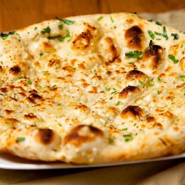 Golden-brown naan bread, topped with garlic and green herbs, rests on a white plate.