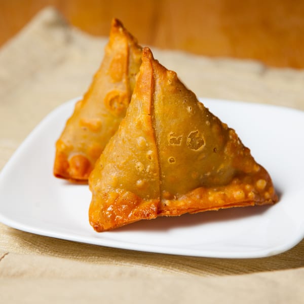 Two golden-brown samosas on a white plate, with small bowls of green and red sauces.
