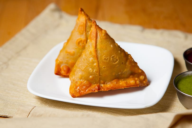 Two golden-brown samosas on a white plate, with small bowls of green and red sauces.